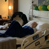 A photo of a bald Anne Sydney with a radiant smile on her face while sitting under a blanket in her hospital bed.