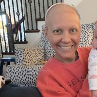 A close-up photo of a bald Anne Sydney smiling while holding up baby Eloise. They are wearing matching white headbands.