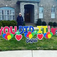 A photo of Anne Sydney smiling while standing in front of her house. There are signs in the yard that read “Congrats,” “Proud of You,” and “You Did It!”