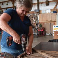 A close-up photo of Charlotte, wearing a navy blue shirt and jeans, using a power drill on a piece of wood in the barn on her farm.