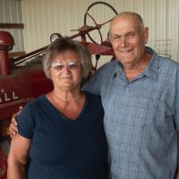 Charlotte and her husband, an older man in a blue button-up shirt, posing for a photo in front of an older red tractor.