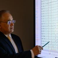Dr. Siddharth Kapoor, pictured in a dark room looking at a projector screen displaying monitor scans while he points at the screen with a pen.