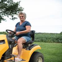 A photo of Charlotte smiling at the camera as she drives a yellow riding lawn mower.