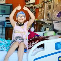 A photo of Ellie in her hospital room where she is sitting upright on the bed with both arms in the air giving two thumbs ups. Her room is full of colorful blankets, stuffed animals, and a yellow gift bag on her bed.