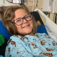 A close-up photo of Ellie laying in her hospital bed smiling at the camera while giving the Star Trek “live long and prosper” hand symbol for the photo. She is wearing a blue gown with cartoon tigers on it.
