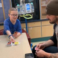 Ellie and Kash sit together at a table in the hospital and play Uno together.