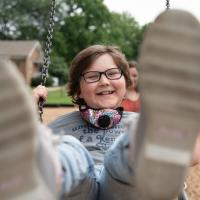 A close-up photo of Ellie swinging and smiling with her feet closest to the camera and blurred slightly. Her mother, Morgan, can be seen in the background.