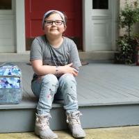 A photo of Ellie sitting on her front porch with her hands on her legs next to her school backpack and lunchbox.