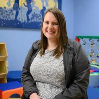 Sarah McAlister, a white adult woman with brown hair, smiles as she sits in the Child Life room at Kentucky Children’s Hospital, surrounded by toys and art. She is wearing a light grey top with a dark grey jacket.