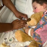Henley and Donnie snuggle in a hospital bed covered by blankets as a nurse checks on Henley’s IV.