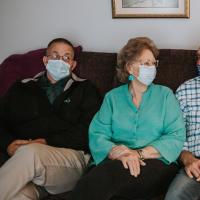 Mae, wearing a turquoise shirt and matching earrings, sits on the couch with three middle-aged men. Each of them are wearing a facemask and are looking at one another.