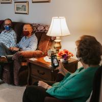 Mae sits on a single couch with her facemask, on a video call with her daughter-in-law, a young white woman with a big smile. Spread across the room are three middle-aged men sitting cross-legged with their facemasks on.