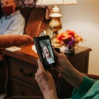A close-up of Mae wearing a mask at home, on a video call with a young white woman who is smiling widely.