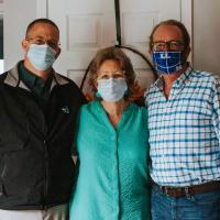 Mae standing closely between her sons. On her left is Kenneth, a middle-aged white man wearing a black jacket, glasses and a facemask. On her right is Scotty, in a white and blue checkered shirt, black framed glasses and a facemask.