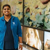 A photo of Dr. Rani Vasireddy smiling while standing in front of a wall decal with sunflowers on it. She is a South Asian woman with long black hair that is tied back in a ponytail. She is wearing a blue UK HealthCare-branded jacket over a pair of dark blue scrubs.