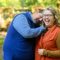 A candid photo of Ronald leaning his head on his wife, Jessica’s, shoulder as she laughs. She is a white woman with long curly blonde hair. She is wearing an orange vest over a long-sleeve brown sweater, and a pair of dark-rimmed glasses.